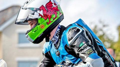 A head and shoulders profile shot of Alex Sinclair on a motorbike looking ahead. His is wearing black and light blue leathers and his helmet is in the colours of the Welsh flag and has the Welsh red dragon on the side.