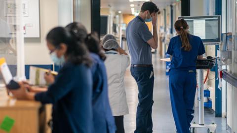 Medical staff in a busy hospital corridor, some looking at a monitor screen.