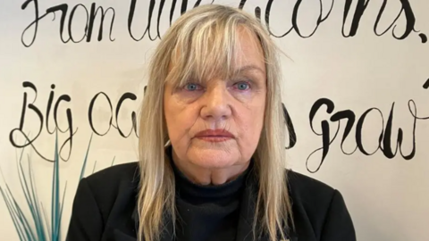 Jean stands for a photo against a white wall. She has blonde hair, with a fringe. She is wearing all black.