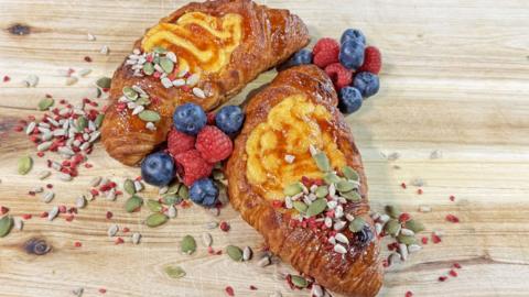 Two Croissants on a wooden board with a crème brûlée filling, dressed with blueberries and raspberries and mixed seeds.
