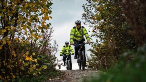 Two police officers in hi-vis jackets and wearing cycling helmets ride e-bikes along a path. Shrubs and trees can be seen to either side of them. 