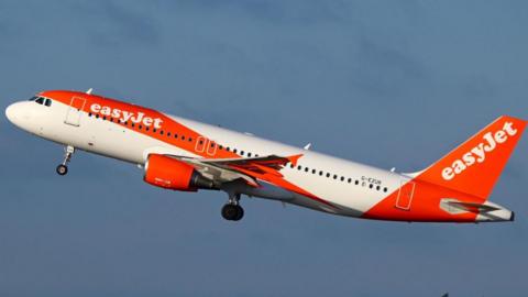 An EasyJet Airbus A320 passenger aircraft climbs after take-off against a clear blue sky. The aircraft is painted in company's distinctive orange-and-white livery.
