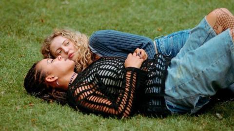 Amelia Clarkson with long curly blond hair wearing a blue top and jeans and lying on the grass in a park. Ebony Aboagye has locs and is wearing a dark-coloured top. She is lying on the grass beside Amelia.