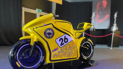 A life-size model yellow bike with the number 26 and text detailing all of Joey Dunlop's 26 wins.
