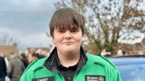 Dylan wearing his green and black St John Ambulance cadet uniform with short brown hair smiling. He is stood in front of trees.