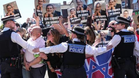 Protesters holding up placards with Keir Starmer's face printed on them being held back by police in Golders Green today 3oth April 2026