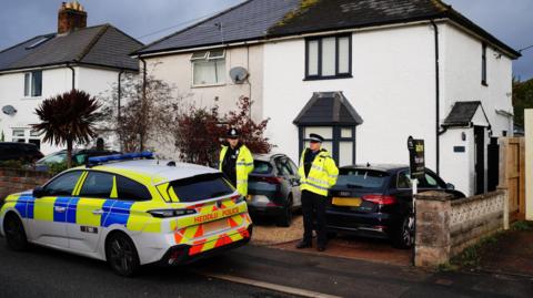 A police car, partially parked on the pavement, outside a white-painted, semi-detached house with black trim on the windows. There is a grey sports car parked on the gravel drive in front of the house, where two police officers in hi-vis yellow jackets stand.