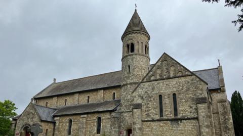 The exterior of St John's church in Hale, a grey building with a tower topped by a turret, built in the Romano architectural style.