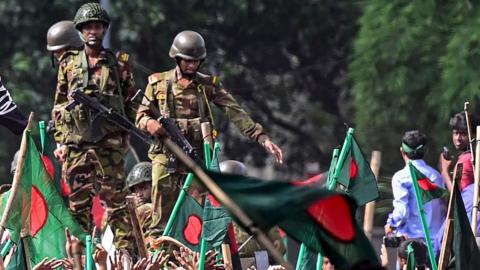 Anti-government protesters wave Bangladesh flags in the foreground while military officers watch over in the background, as the protesters march towards the prime minister's palace in Dhaka, Bangladesh on 5 August