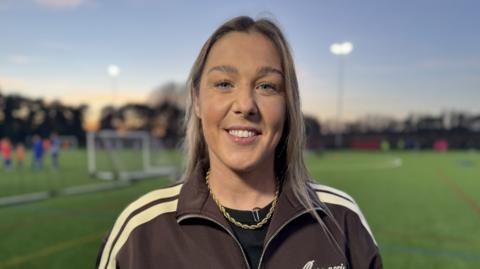 Mary Earps smiles as she looks into the camera and stands on the new football pitch. She's wearing a black top underneath a brown jacket with cream stripes down the shoulders as well as a gold necklace.
