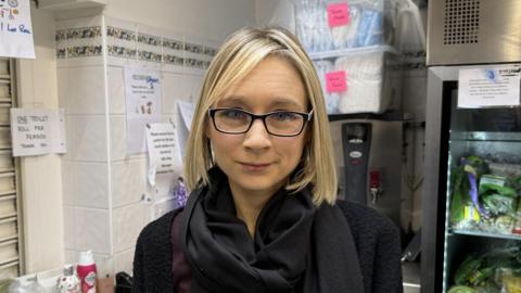 A woman with blonde hair wearing glasses and a black scarf, stood in front of a fridge. 