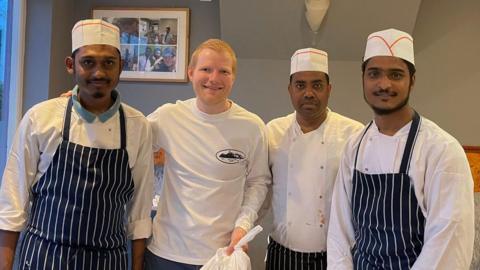 Three men wearing chef whites and hats as well as blue aprons stand with another man with red hair wearing a white long sleeved T-shirt who is holding a white bag filled with a food order. 