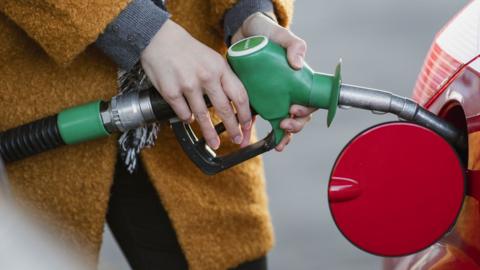 Close up of someone filling up a red car with petrol from a petrol pump. The person is wearing a mustard coloured coat. All you can see is their hand on the pump.