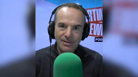 Martin Lewis wears a black top and headphones and sits in front of a green microphone