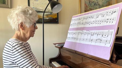 A woman sits in front of a piano reading large sheet music as she plays. She has short grey hair and wears a white long sleeved top with black stripes on it. She is also wearing glasses. 