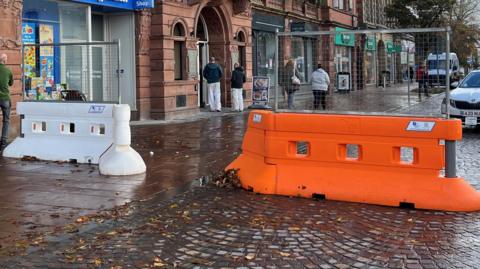 The entrance to the pedestrian area by the Crown and Mitre in Carlisle, which is a large and beautiful redbrick building with leaded windows, a first floor balcony running most of its length, ornate stone work and many chimneys. The ground floor is given over to shop fronts. A taxi is parked behind a large, plastic temporary orange barrier, next to which is a white barrier of the same kind just outside a toy shop.