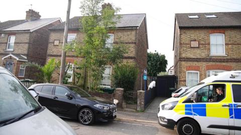 Image shows a semi detached brick house with a tree in the front garden. There is a car and a police van parked outside.