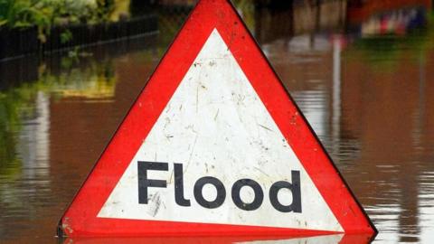 A red triangular road sign warning drivers of a flood. It is half submerged in murky brown water. In the background there is a hedgerow on the left.