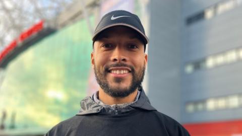 Joe Thompson, wearing a baseball cap, smiles in front of Manchester United's stadium