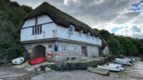 Coronation Boathouse at Bantham, a thatched two-storey boathouse in a mock Tudor style with a number of tenders tied to a harbour wall in front of it on a pebbly foreshore.