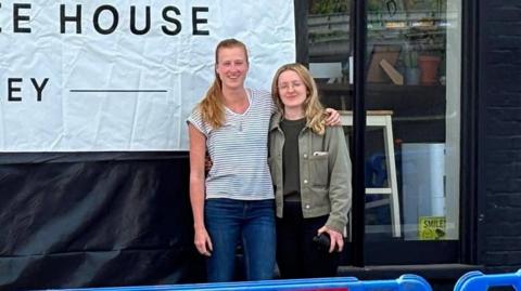 A blonde woman in a white striped t-shirt and jeans, standing next to a blonde woman in a green jacket and black trousers. They are standing in front of a shop and there is a large white banner which reads The Coffee House Nutley.