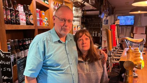 Trevor and Julie stood behind the bar in their pub. They are in front of the lager taps and there is bottles of beer and cans of soft drinks on the shelves.