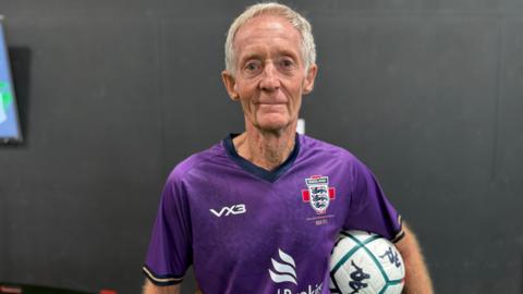 Photo of a man with short grey hair looking into the camera. He is wearing a purple football shirt with an England badge on the right. He is also holding a while football with turquoise detailing.