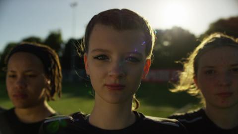 three women stare at the camera while standing on a grass pitch with the sun behind them