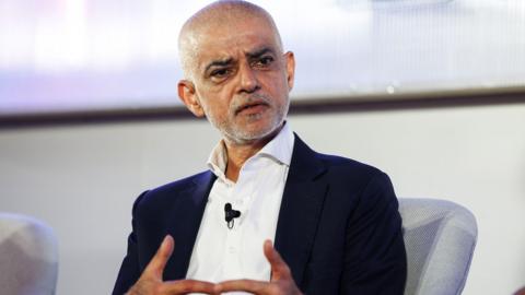 Sadiq Khan sitting in a light grey chair wearing a dark suit and a white shirt. 
