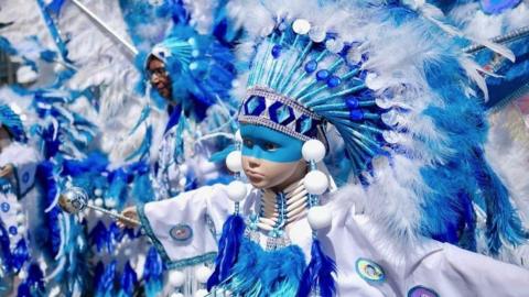 A very striking image of a child dressed in a blue and white feathered outfit, with a large blue and white feathered headdress and blue paint across the top part of their face. Their arms are outstretched and there are people in the background wearing similar outfits.