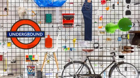 A range of everyday objects include as pair of jeans, a rec hot water bottle, a red circular sign with the word Underground in white running across the centre. There is also a black bicycle, a mop with a string head and a vibrant green table.