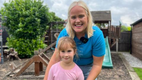 Emily is wearing a pink t-shirt and standing in a playpark in front of her mum. There's a wooden seesaw and a playhouse with a slide behind them. 