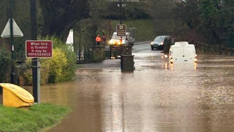 A white van can be seen driving through water on a flooded road, while other vehicles are stopped in the distance beyond the flood. There is a road sign which says "when road is flooded it may be impassable".