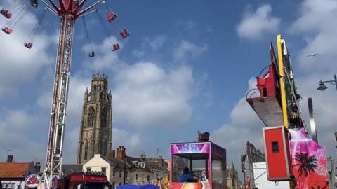 A high swing ride is on the left of the photo with a church and houses in the middle. A booth for a ride is in the middle of the image and a person is sitting inside.