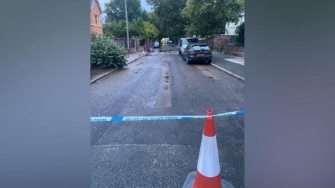 A traffic cone and police tape can be seen at one side of a road, with some water seen on the road and an emergency services vehicle at the other end of the road.