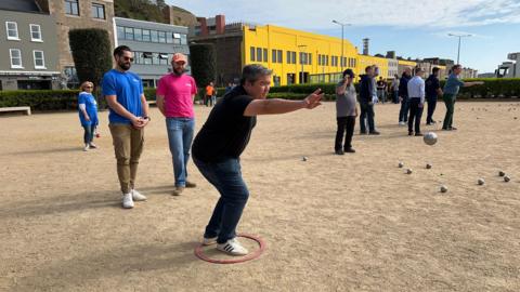 A man stands crouching in a red ring after he has thrown a boule as two men watch him and in the background more teams are playing pétanque 