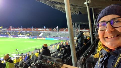 John Murray smiling with the backdrop of the ground at Bodo/Glimt's Champions League game against Manchester City