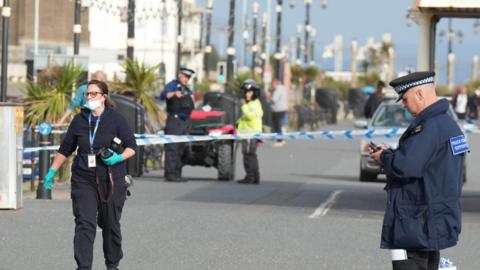 A number of police officers, including a forensics officer holding a camera, on Worthing seafront following a stabbing in the area. A cordon can be seen in the background of the image.