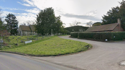 A google maps image of St. Nicholas's Church in Pyrford, near Woking, Surrey. part of the church can be seen in the image alongside an entrance and a grassy area.