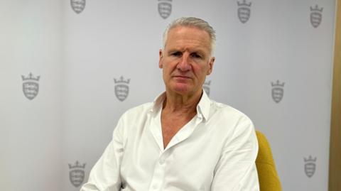 Jersey Health and Social Services Minister Tom Binet sits on a yellow chair in front of a board with the Government of Jersey's logo on it. He is wearing a white shirt with the top couple of buttons undone.