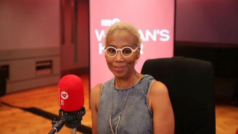 Trisha Goddard sits in front of a red Woman's Hour microphone. She is wearing large white glasses and a denim top.