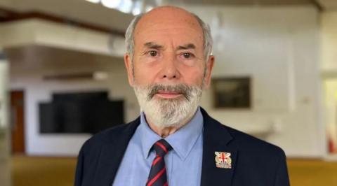 A man with a grey beard in a blue suit and a stripped tie.