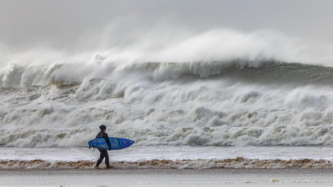 The picture shows a lone surfer standing on the shoreline, holding a blue surfboard and looking out towards huge, churning waves. The sea is rough and wild, with towering white‑topped swells crashing in the distance. The sky looks grey and stormy.