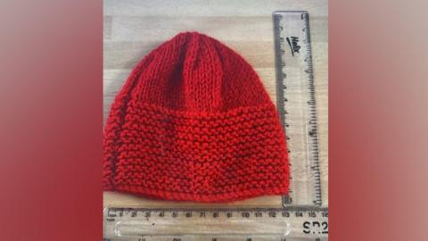 Red knitted hat with contrasting patterned edge alongside two small plastic rulers which indicate the size. 