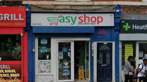 General view of a row of shops. In the centre of the photo is a  white and blue store named Easy Shop, the E of of the Easy is a shopping trolley.