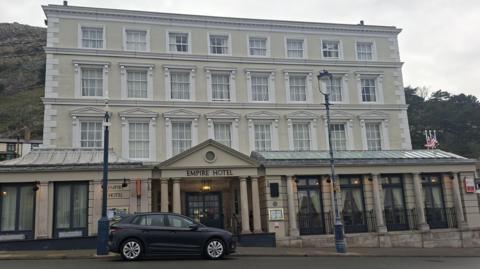 The Empire Hotel in Llandudno - it is a cream building with white windows standing three storeys tall. On the street in front of the building is a black car and the Great Orme rises behind the building in the background