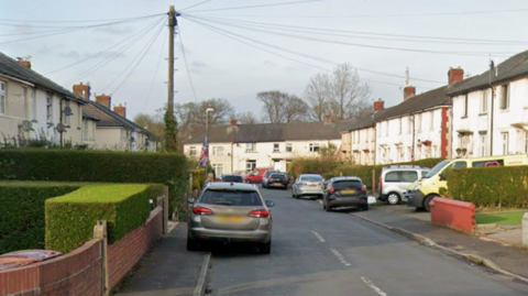 Google street view showing gardens and white houses lining the road with cars parked on both sides.