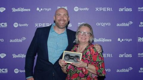 Joe Sims, a bald man wearing a blue shirt and dark jacket, stands smiling next to Nicki Ledgard, a woman with blonde hair and glasses, who is holding a trophy that looks like an audio speaker.