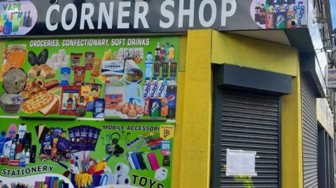 Image shows the brightly coloured exterior of The Corner Shop in Picton Road, Liverpool. Advertisements for vapes, stationery, toys and sweets adorn one of its walls. The shutters are down and there is an enforcement notice taped to them. 