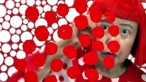 Yayoi Kusama seen looking through a clear screen with red spots painted on it, with red and white clothes and background